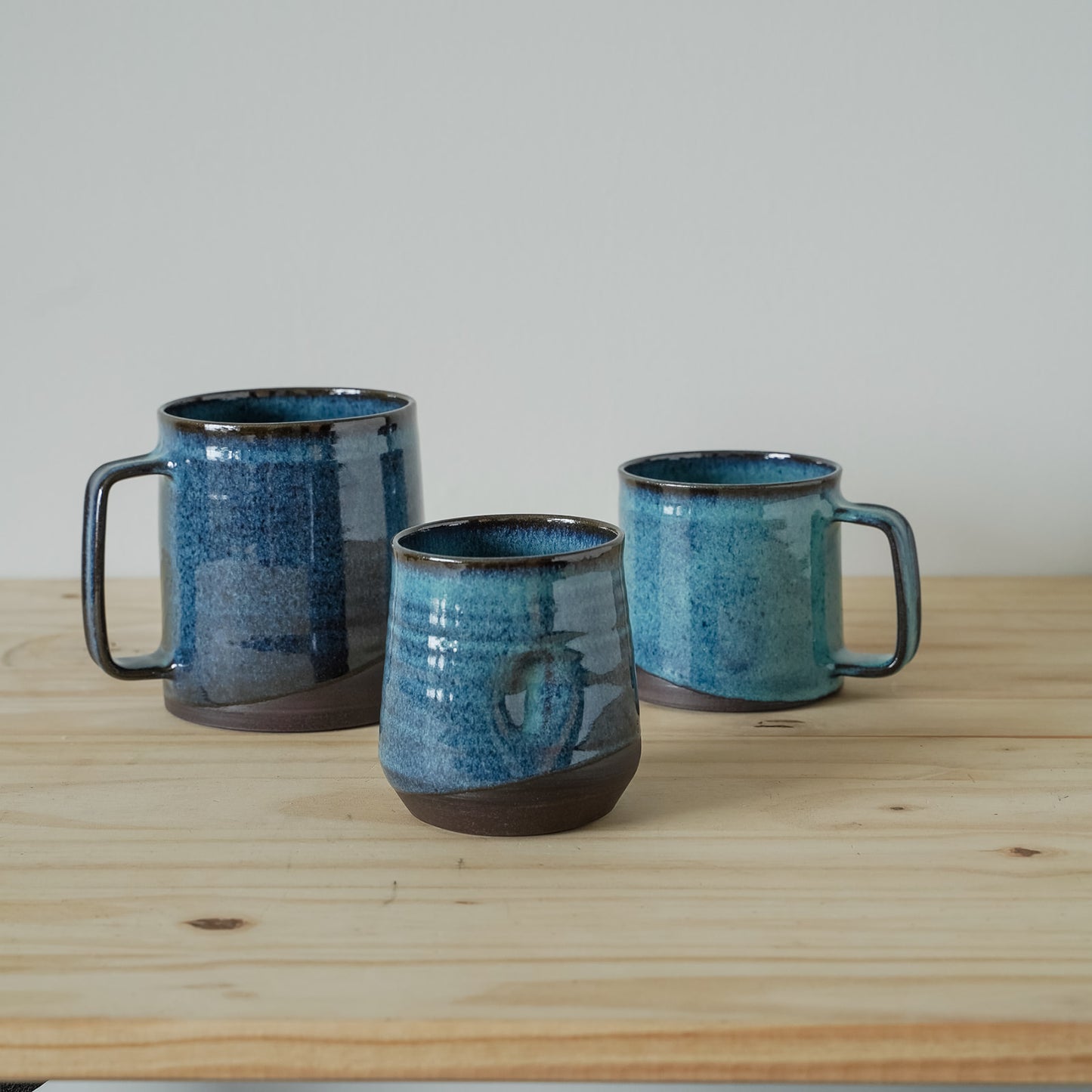 Three glossy Sea Glass ceramic mugs on a wooden table with a neutral background. Two mugs have handles, and one does not.