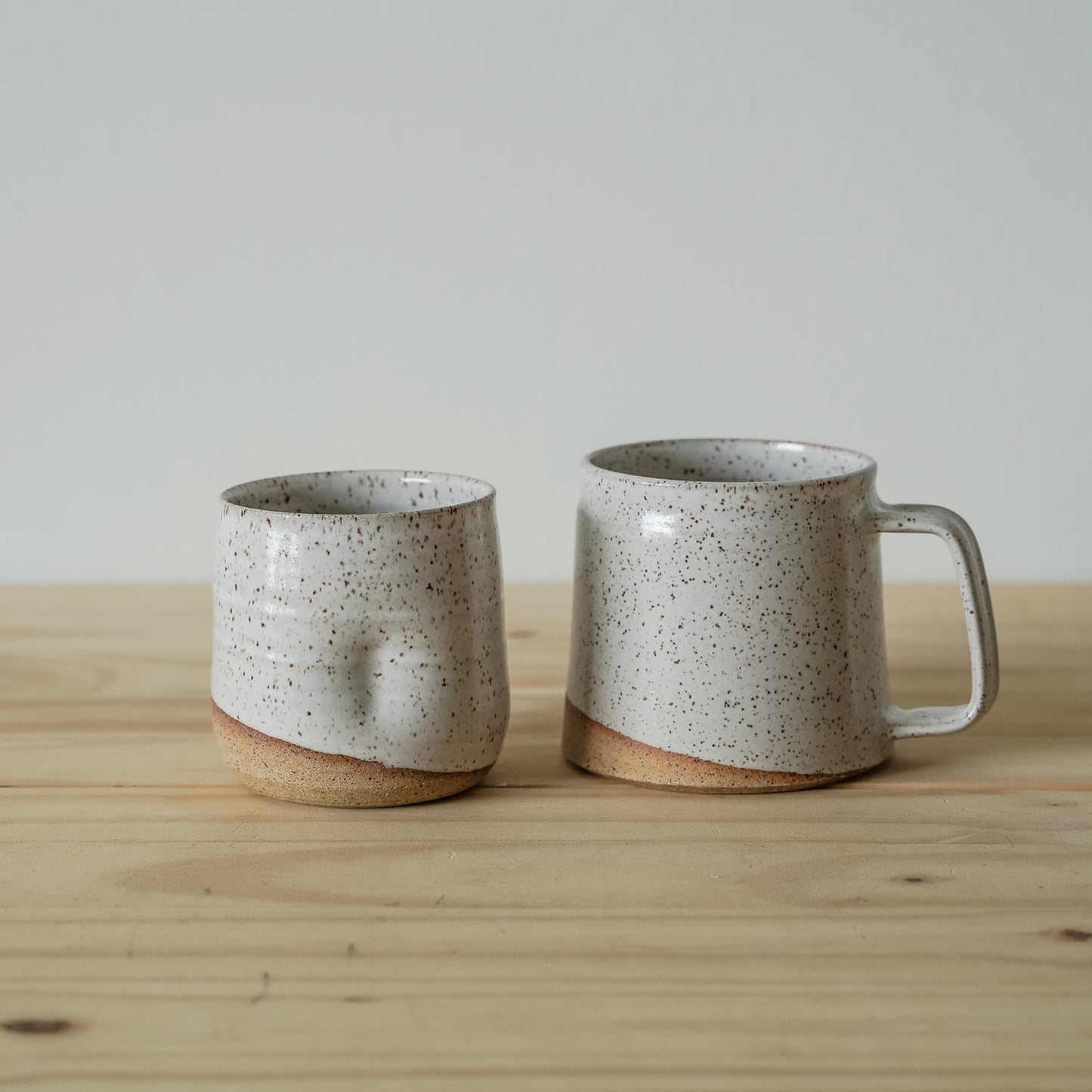 A speckled white ceramic tumbler and mini-barrel mug with earthy tones sit on a wooden surface creating a handmade aesthetic against a plain backdrop.