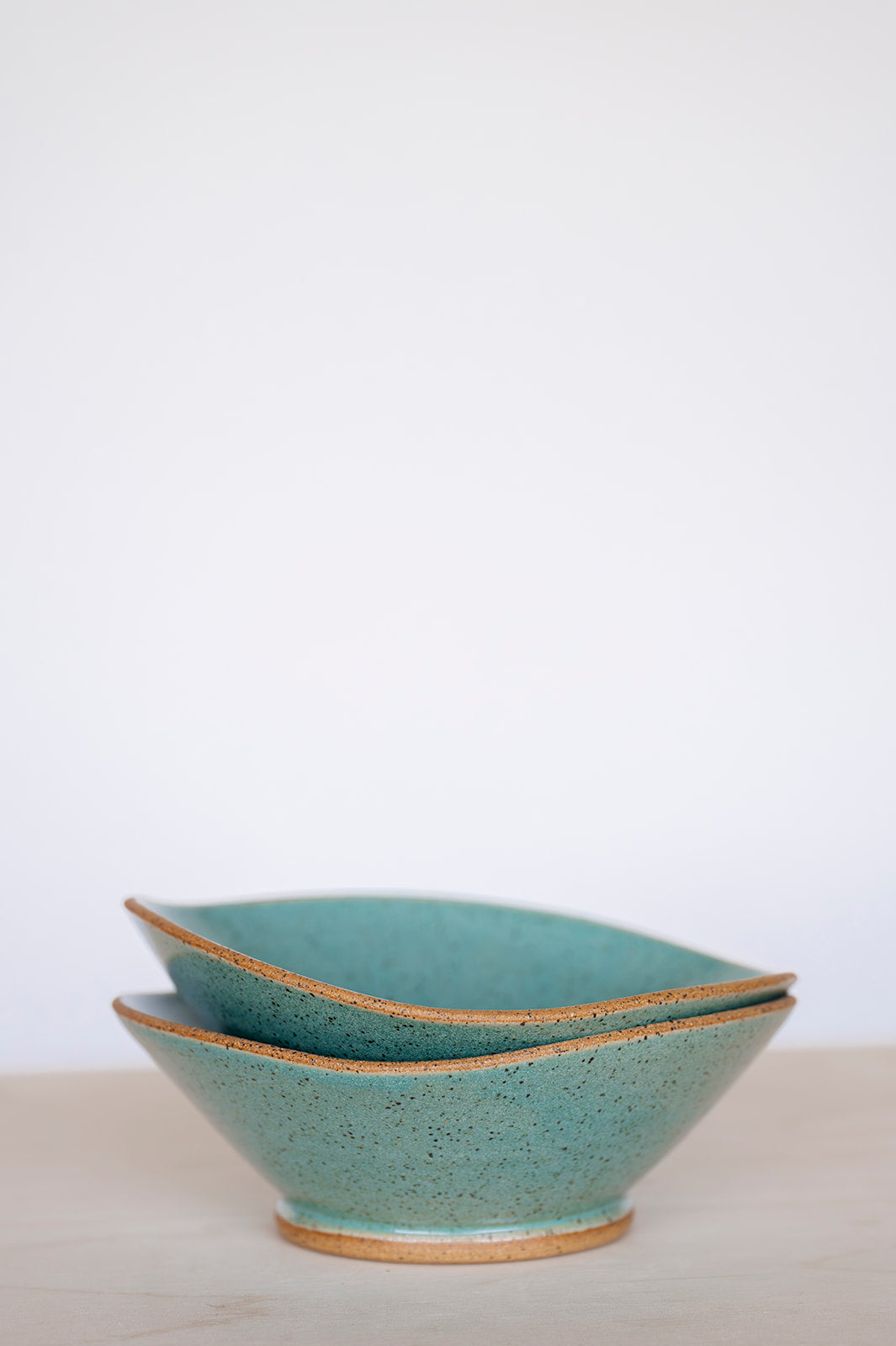 Two stacked, handcrafted ceramic bowls with speckled jaded glaze and raw tan edges, set against a soft, neutral background, conveying a serene and minimalist tone.