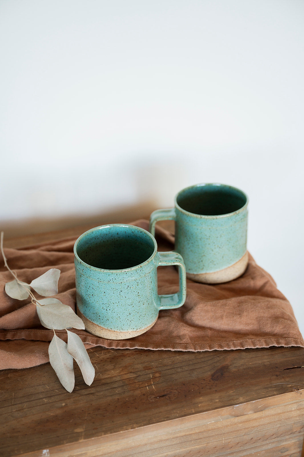 Two handmade speckled jaded mugs sit on a oragne/brown cloth on a wooden table, accompanied by pale, dry leaves. The scene conveysa rustic, calm vibe.