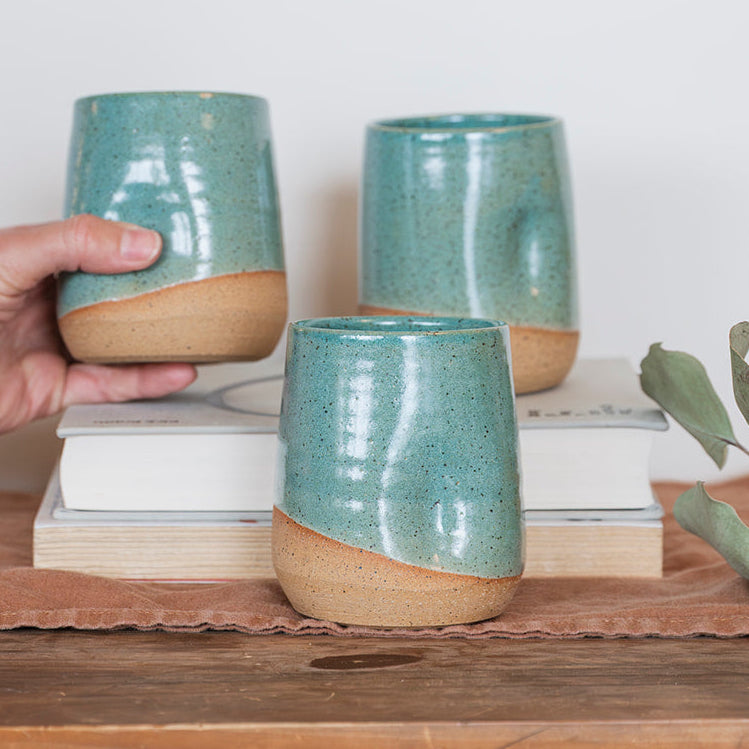 A set of three handmade 12 ounce Speckle Jaded glaze ceramic tumblers. A hand holding one in the background on the left with the thumb resting in grip groove.