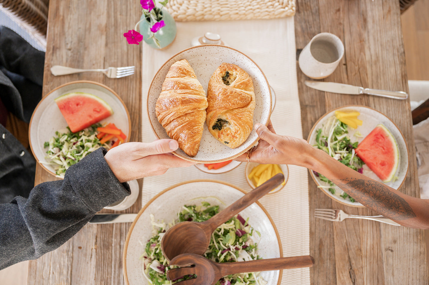 Two people share a plate of croissants over a rustic wooden table set with salads, watermelon, and flowers, creating a cozy brunch atmosphere using Good Wheel Ceramics hand crafted dinnerware.