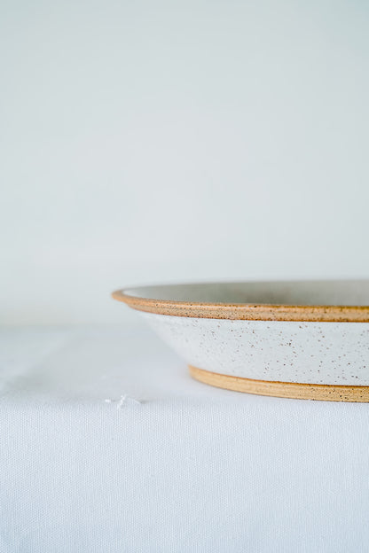 A speckled white Good Wheel Ceramics serving bowl with a contrasting brown raw rim sits on a white tablecloth against a pale background, conveying a minimalist and serene tone.