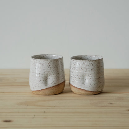 Two rustic ceramic tumblers with a speckled white glaze sit on a wooden table. Their uneven texture and earthy tones evoke a handmade, cozy feel.
