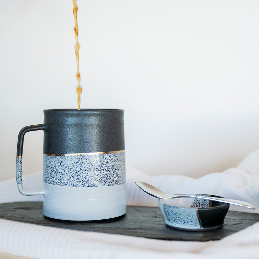 Coffee being poured into a black, white and gold ceramic mug on a wooden surface with a white background