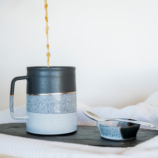 Coffee being poured into a black, white and gold ceramic mug on a wooden surface with a white background