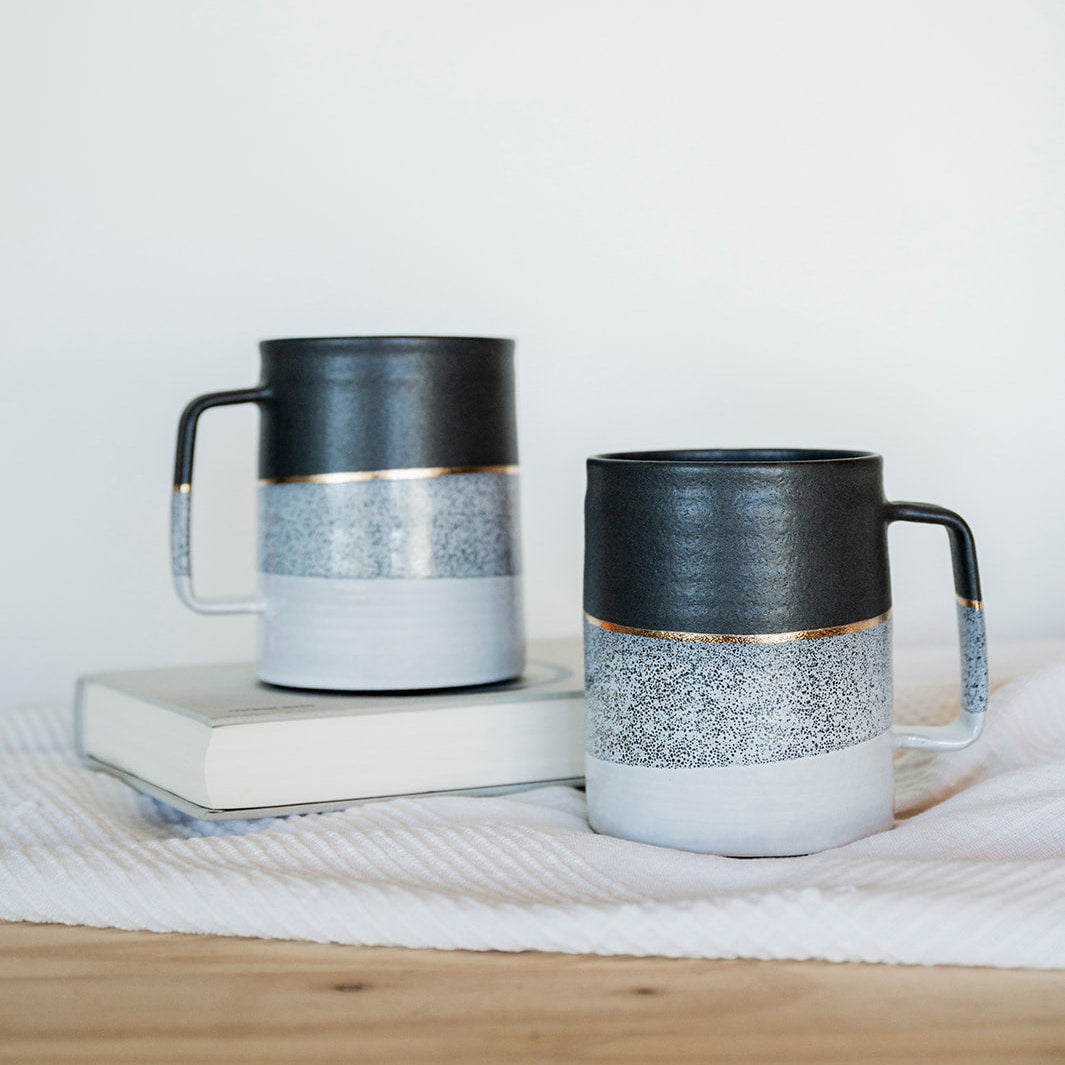 Two ceramic mugs with black and white and gold design on a wooden surface with a white background
