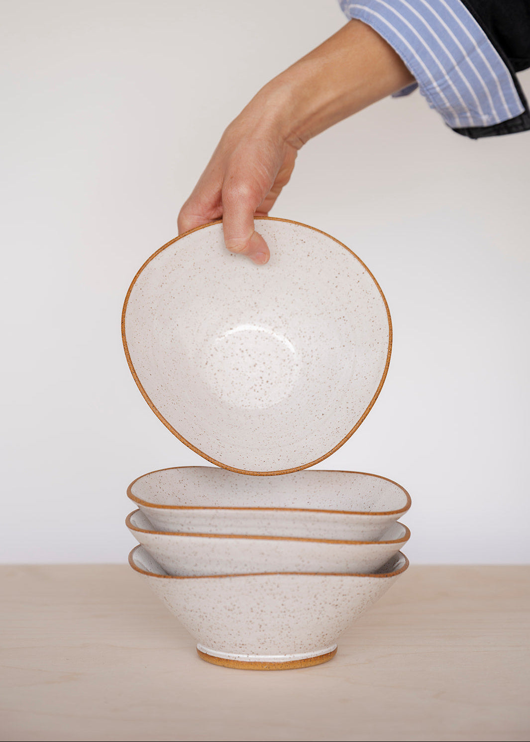 A hand in a blue-striped shirt sleeve holds a speckled white Triangle bowl above a stack of three similar bowls on a light wooden table.