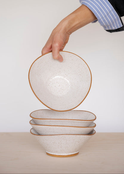 A hand in a blue-striped shirt sleeve holds a speckled white Triangle bowl above a stack of three similar bowls on a light wooden table.