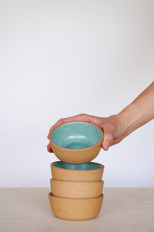 A hand holding a Speckled Jaded Good Wheel Ceramics Snack bowl above a stack with a plain background. 