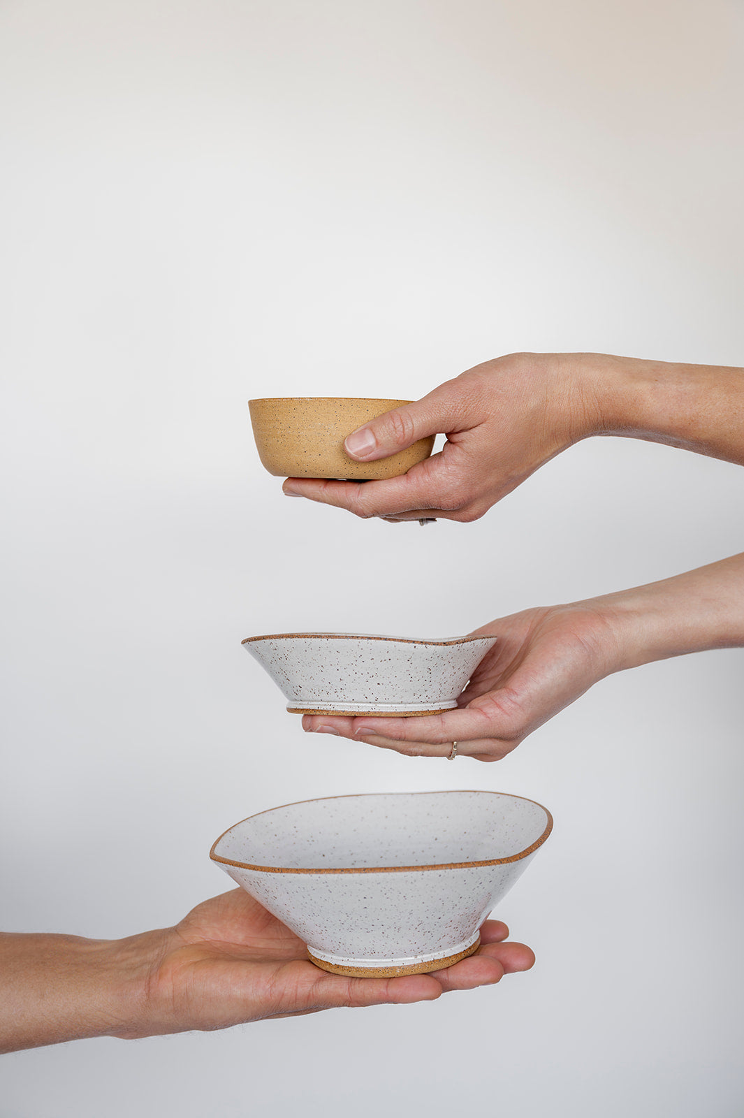 Three hands hold three uniquely shaped Good Wheel Ceramics bowls against a white background. The top Snack bowl shows the raw clay outside, while the middle and bottom are glazed white with a contrasting raw rim. The arrangement is minimalistic.