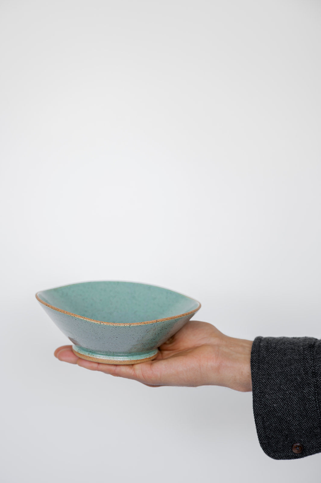 A hand holds a small, speckle jaded ceramic bowl with a satin matte finish against a plain white background. The scene conveys simplicity and elegance.