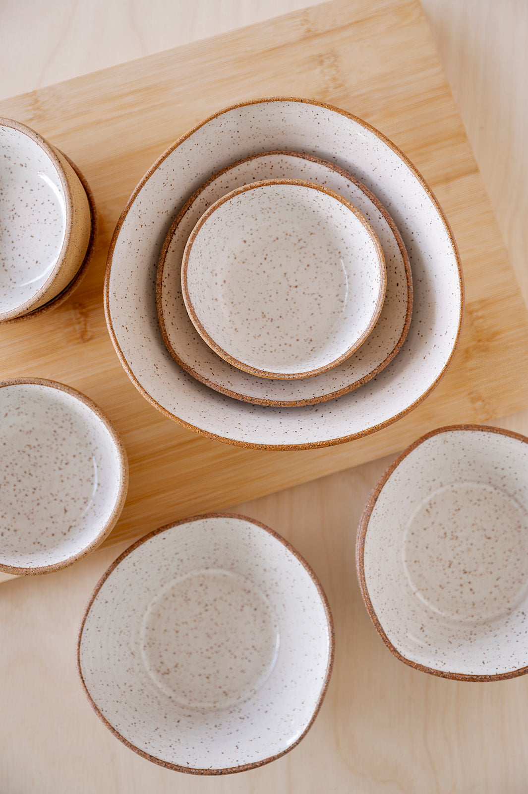 A set of speckled white Good Wheel Ceramics bowls with brown rims arranged on a wooden surface. The bowls are nested and scattered, creating a rustic, warm feel.