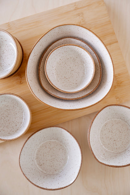A set of speckled white Good Wheel Ceramics bowls with brown rims arranged on a wooden surface. The bowls are nested and scattered, creating a rustic, warm feel.