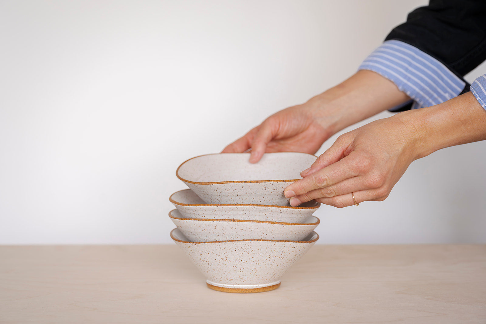 Two hands arranging a stack of four speckled white ceramic Triangle bowls on a wooden surface. The scene conveys a sense of calm and careful organization.