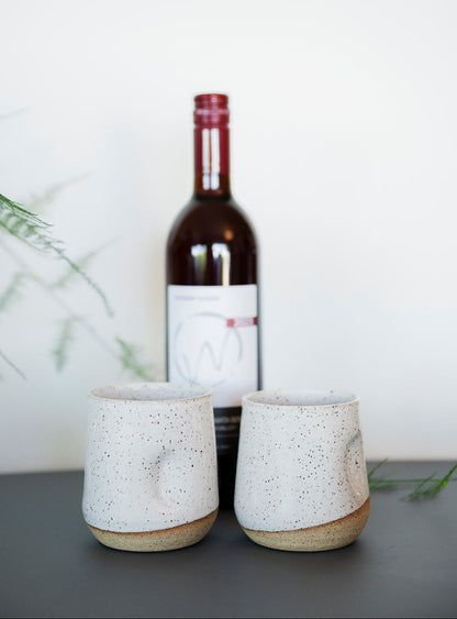 A bottle of red wine with a white label is positioned behind two speckled white tumblers on a gray table, surrounded by soft green foliage.