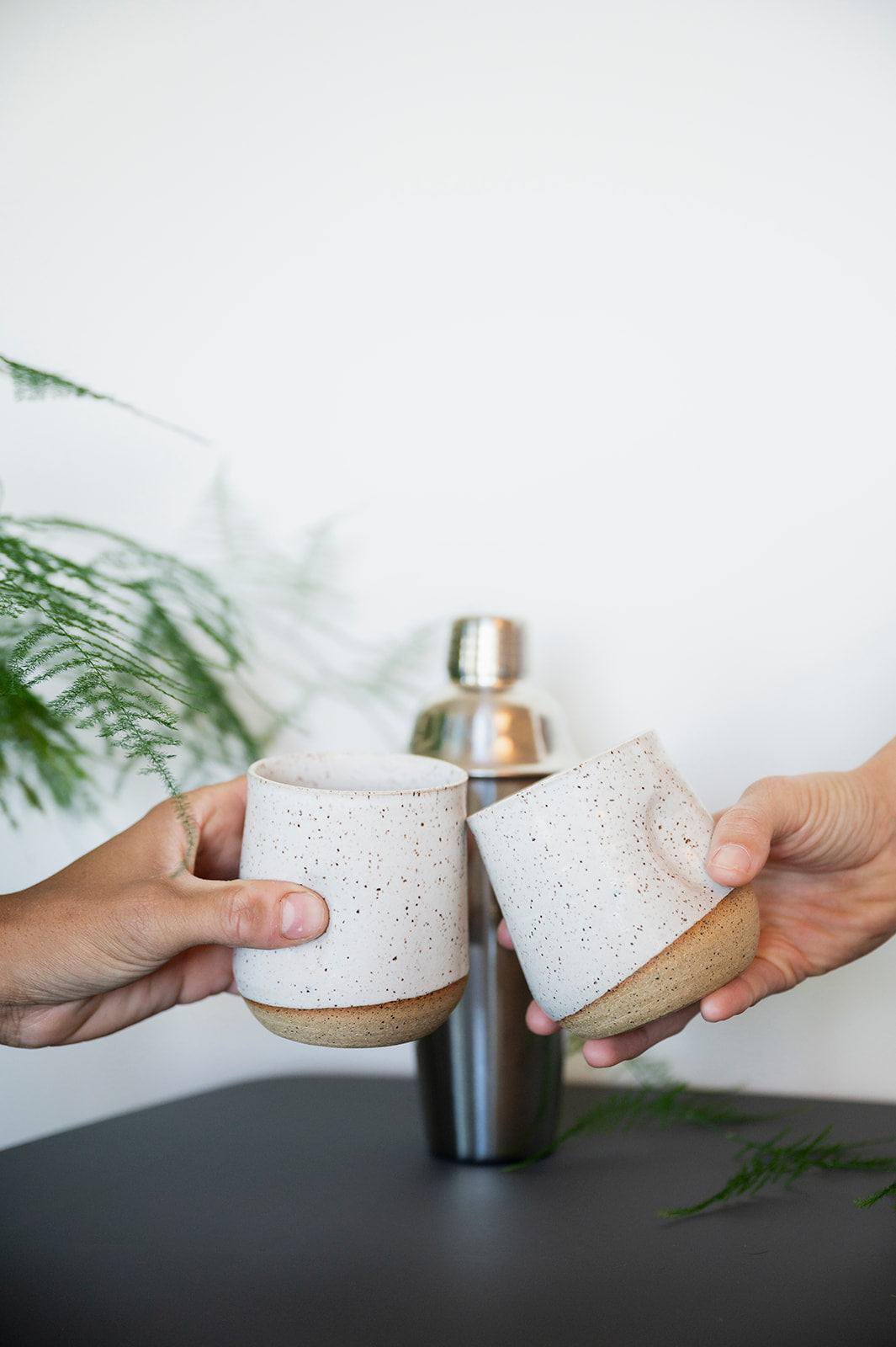 Two hands toast speckled white tumblers against a minimal background with greenery. A metal cocktail shaker sits on the table, conveying a casual, festive vibe.