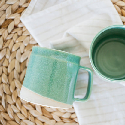 Two green ceramic mugs 

Handmade ceramic mini barrel mug in Jaded glaze styled on a woven surface with a white cloth underneath. Artisan stoneware mug showing glaze texture and size, small-batch pottery British Columbia