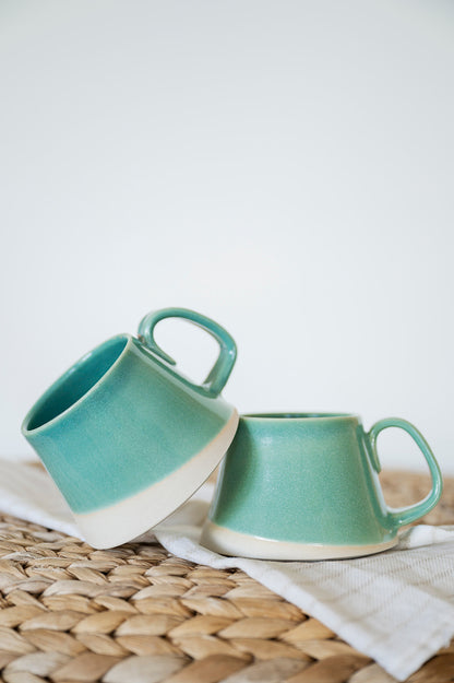 Two handmade jaded ceramic A-frame mugs on a woven mat with a white background. Comfortable two-finger handle, wheel-thrown on Vancouver Island, Canada. 