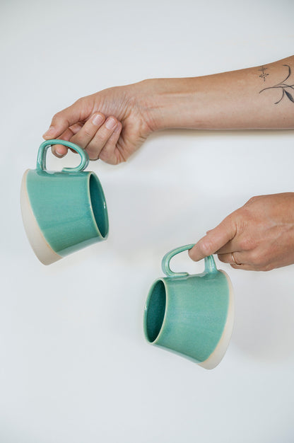 Two hands dangling handmade jaded A-frame mugs by the handles against a white background, showing the unique shape and taper. 