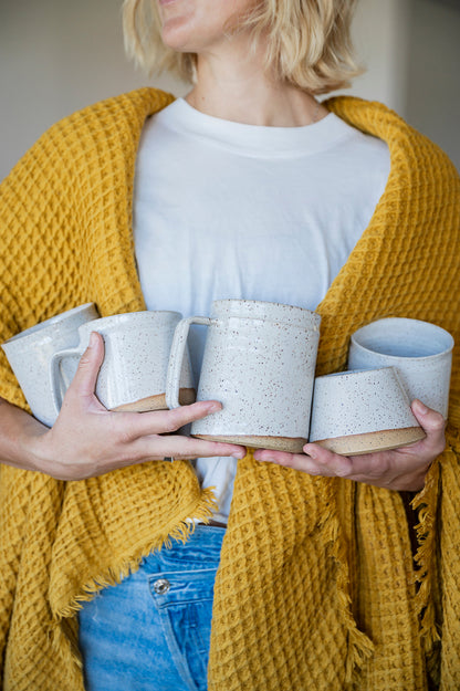 A woman wearing a white shirt with a mustard yellow waffle blanket draped over her shoulders holding 5 speckled white Good Wheel Ceramics mugs in her arms. Handmade on Vancouver Island. 