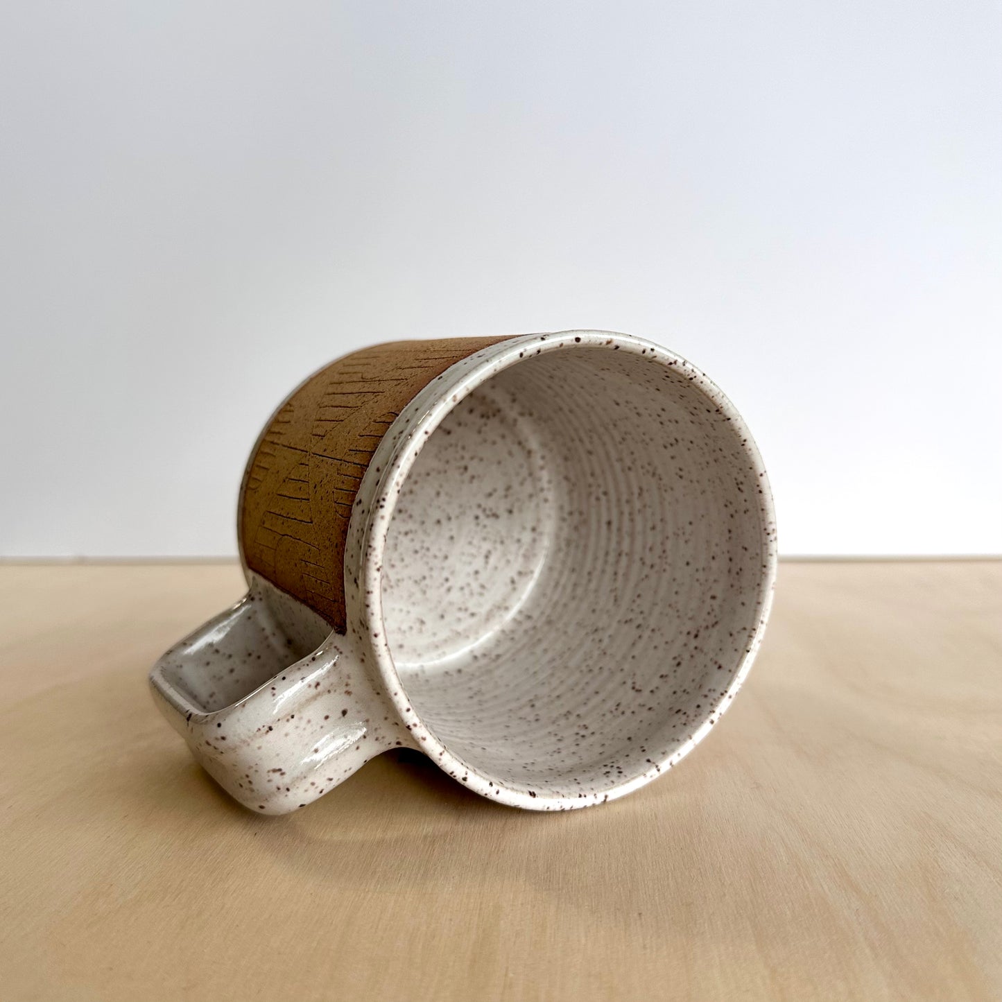 Ceramic mug with hand-carved geometric design sitting on its side on a wooden surface showing the inside white glaze. 