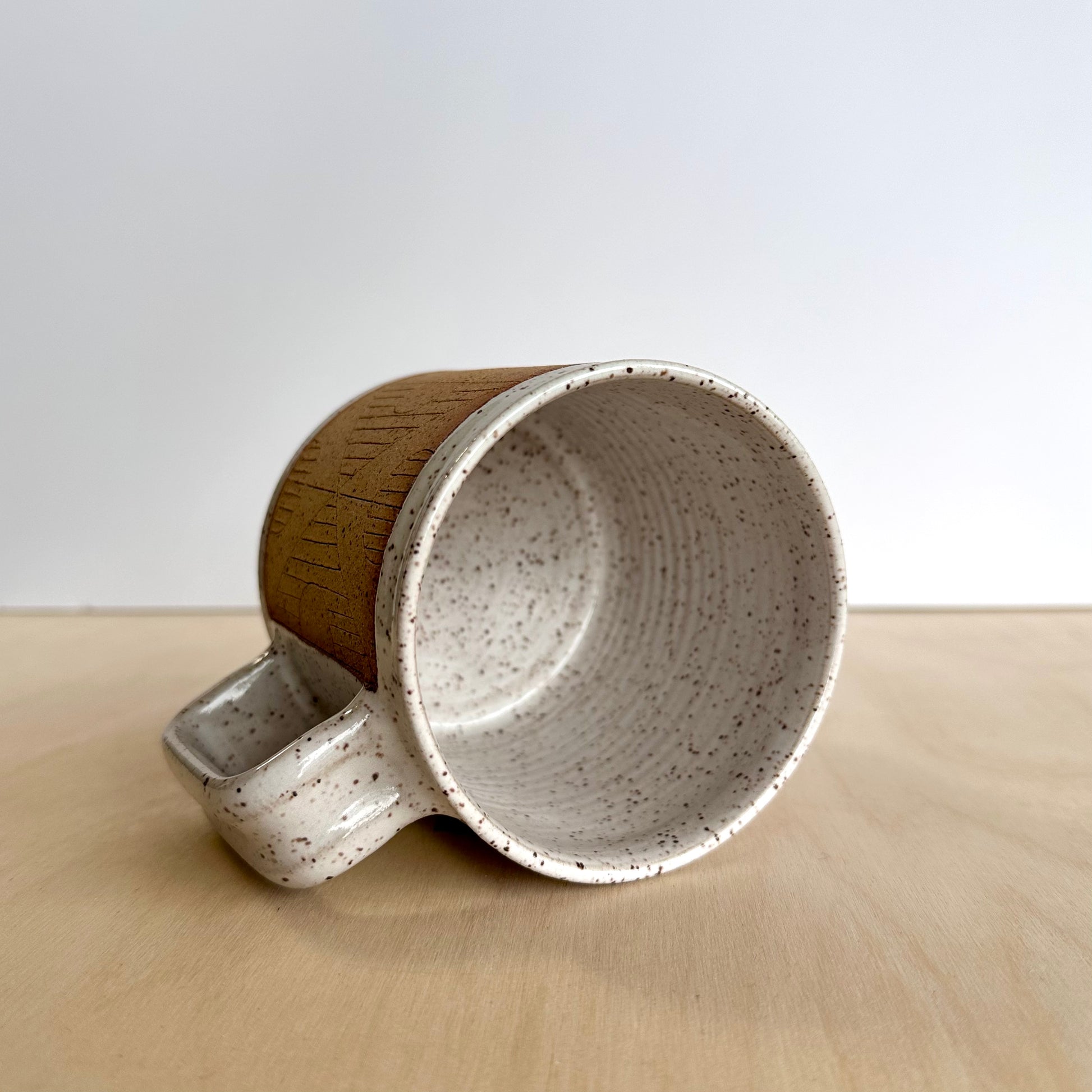 Ceramic mug with hand-carved geometric design sitting on its side on a wooden surface showing the inside white glaze. 