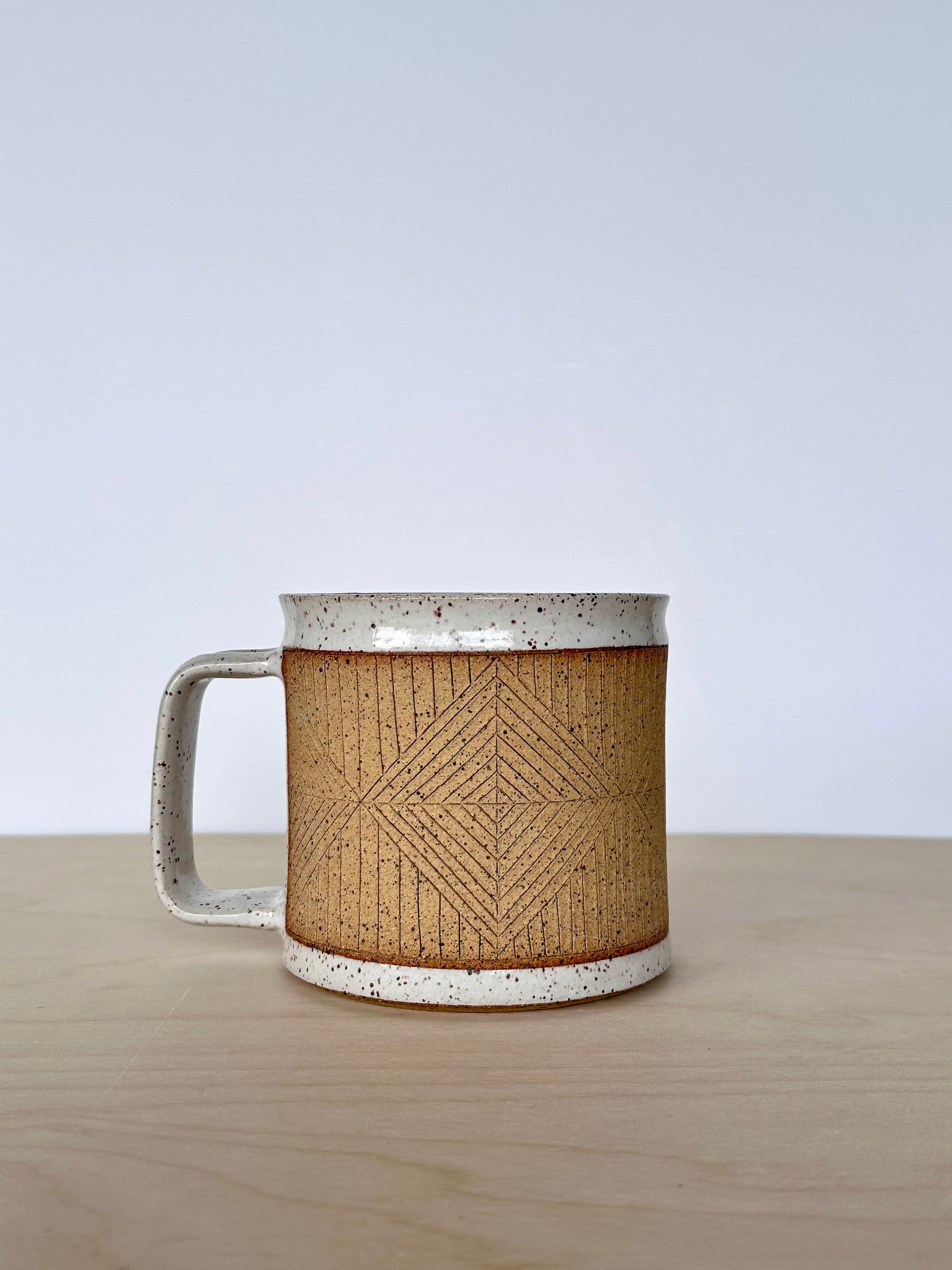 Ceramic mug with hand-carved geometric design sitting on a wooden surface, handle facing left. 