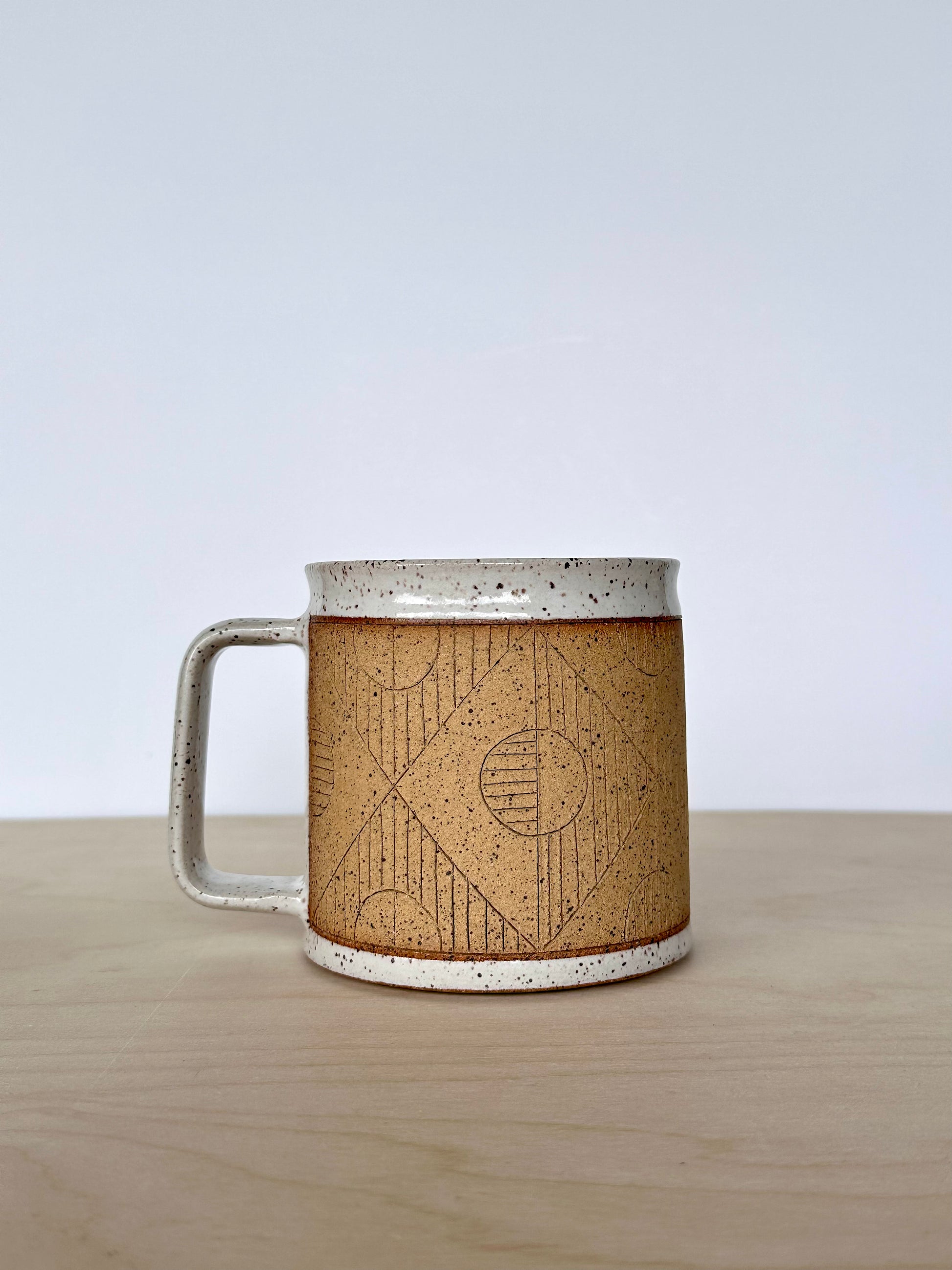 Ceramic mug with unique circles and lines hand-carved geometric design sitting on a wooden surface, handle facing left. 