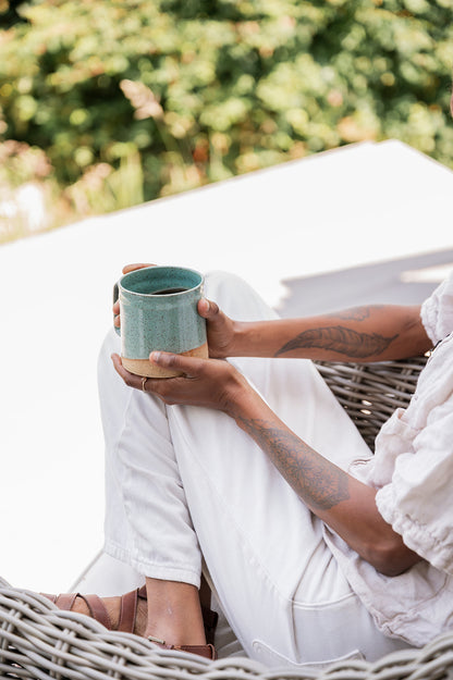 A person wearing white pants sits in a wicker chair, holding a handmade 16oz speckle jaded mini-barrel ceramic mug. Sunlit greenery and a relaxed atmosphere are in the background.