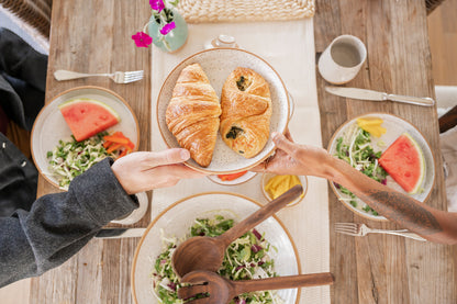 Two people share a plate of croissants over a rustic wooden table set with salads, watermelon, and flowers, creating a cozy brunch atmosphere using Good Wheel Ceramics hand crafted dinnerware. 