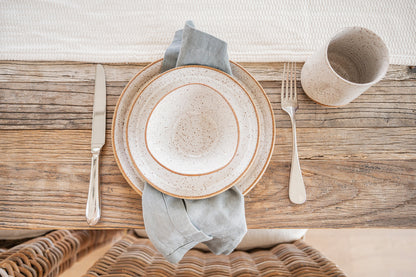 A rustic table setting features Good Wheel Ceramics speckled white dishes and tumbler on a weathered wooden table. A blue napkin, silver cutlery, and a woven chair complete the cozy, homely vibe.