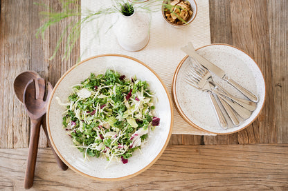 A fresh green salad in a large Good Wheel Ceramics serving bowl, set on a rustic wooden table with wooden salad servers, a small vase, cutlery on a stack of dinner plates, and a Snack bowl with chopped mushrooms. 