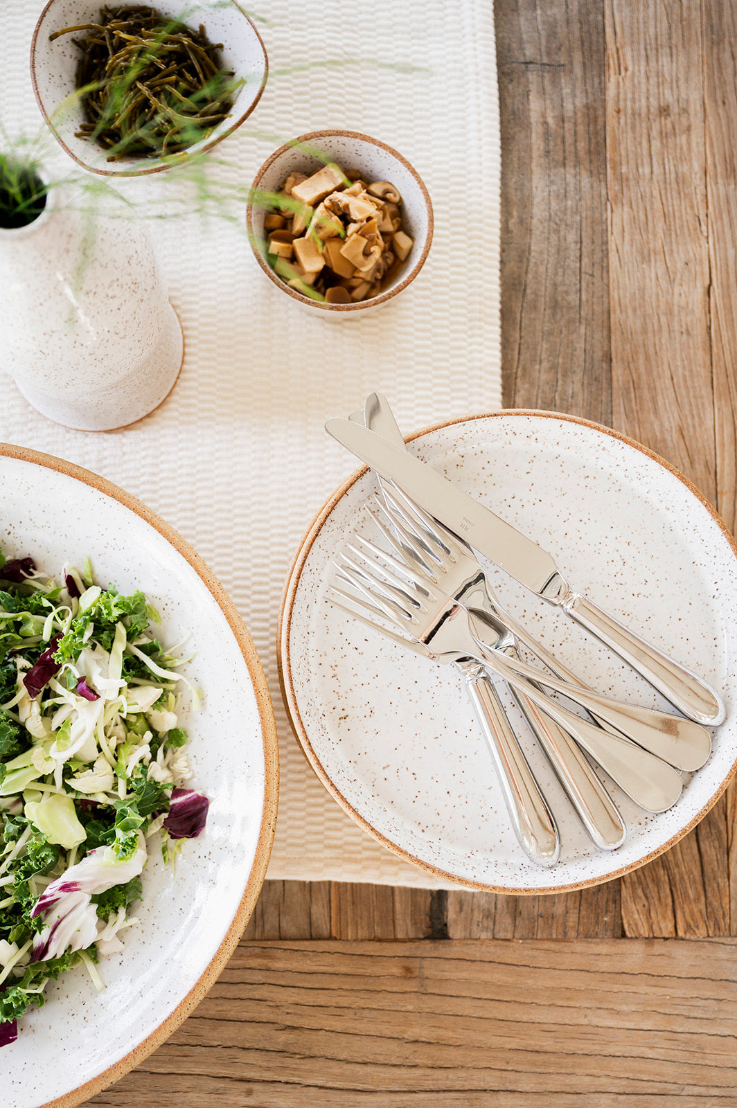Table setting with of Good Wheel Ceramics dishes cutlery, a bowl of salad, and small snack bowls on a wooden table.