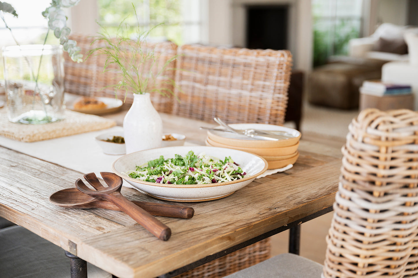 A rustic wooden table set for a meal with a fresh salad in a Good Wheel Ceramics serving bowl, wooden utensils, white vase and dinner plates. Wicker chairs add a cozy, inviting feel.