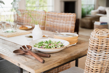 A rustic wooden table set for a meal with a fresh salad in a Good Wheel Ceramics serving bowl, wooden utensils, white vase and dinner plates. Wicker chairs add a cozy, inviting feel.