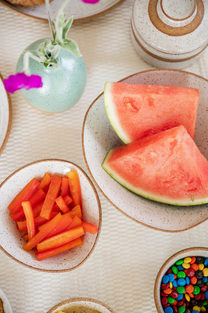 Ceramic Triangle, Mini-triangle and Snack bowls on a textured tablecloth hold watermelon slices, bright red pepper sticks, and colorful M&Ms. A small speckle jaded vase with pink flowers adds a cheerful touch.