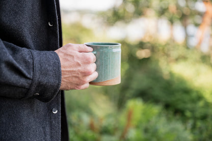 Close-up of a person wearing a dark jacket holding a speckle jaded mini-barrel mugs ceramic mug outdoors. Background is blurred with lush greenery, conveying a serene atmosphere of enjoying a morning coffee outdoors. 