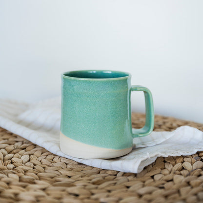 A single handmade Jaded glaze ceramic mini barrel mug, three-finger handle stoneware mug 16 oz, wheel-thrown pottery Vancouver Island Canada, sitting on a woven texture. 