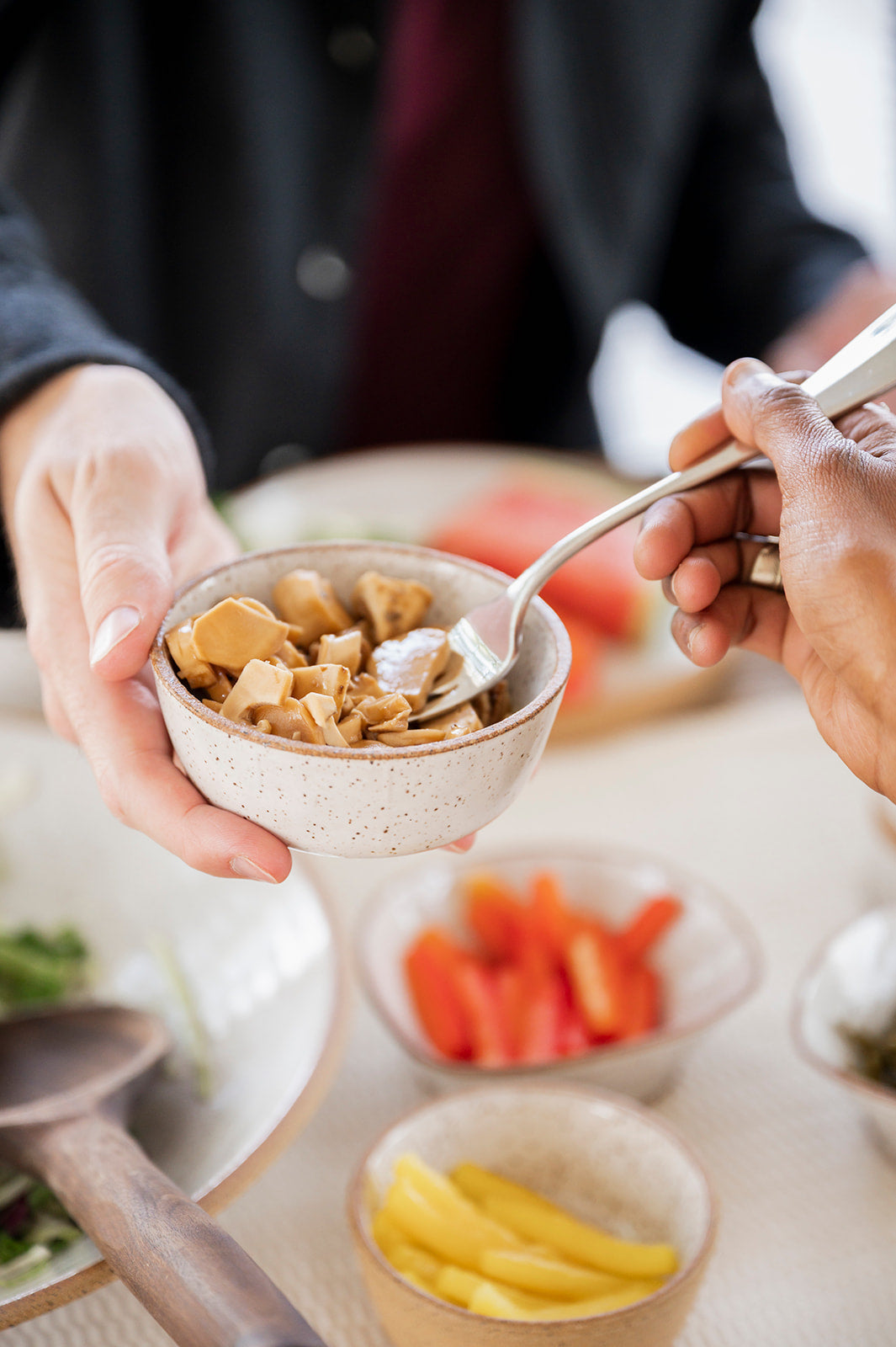 One person holding a bowl while another person uses a fork to serve themselves from it. Surrounded by various food items on the table in the background.